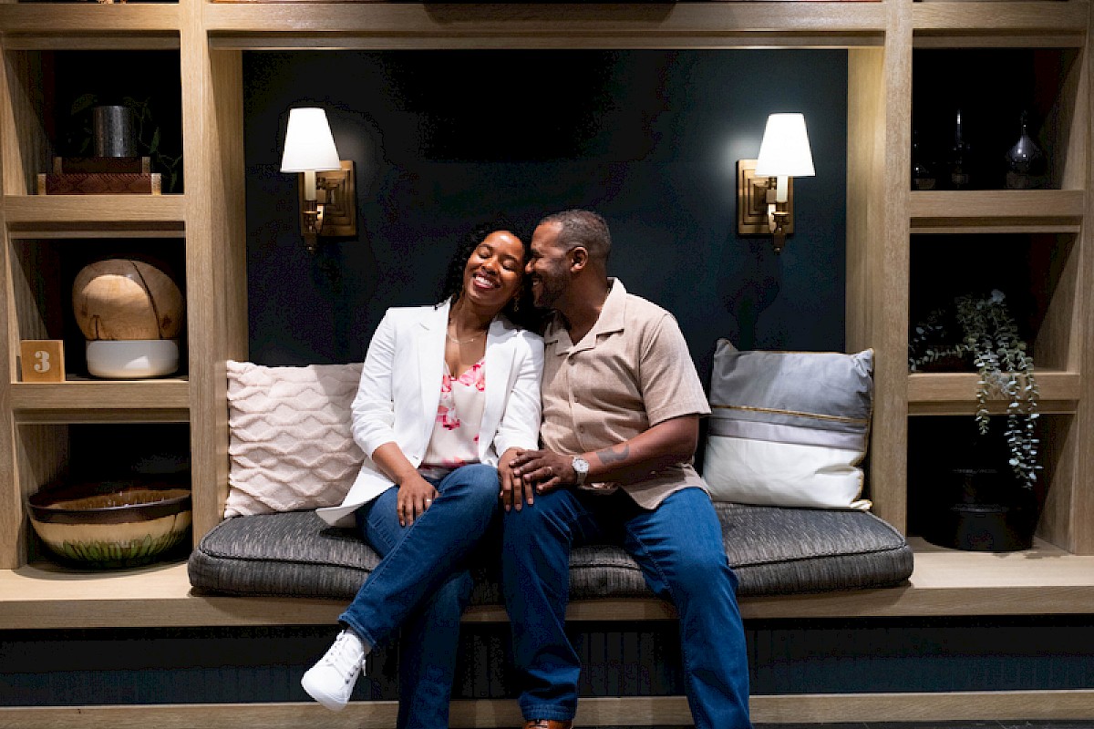 A couple sits closely on a bench in a cozy, stylish room with shelves, cushions, and warm lighting.