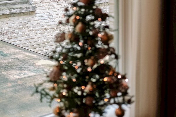 A small, festive Christmas tree on a windowsill with warm lights and ornaments, alongside a round wooden tray holding two candles by the window.