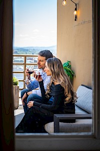 A couple sits on a balcony, enjoying glasses of wine with a scenic view in the background, captured through a doorway.