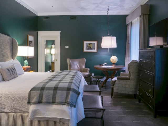 A cozy bedroom with a double bed, chairs, a small round table, a dresser, and a TV, all lit warmly with lamps and natural light.