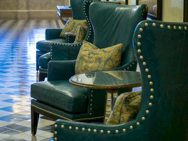 The image shows a row of comfortable green chairs with decorative pillows and small round glass-top tables on a tiled floor.