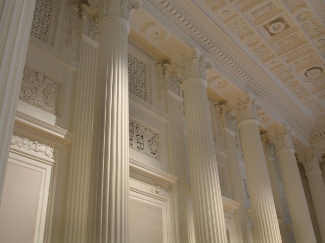 The image shows an interior view of a building with tall, ornate columns and detailed architectural molding and decorations on the walls and ceiling.