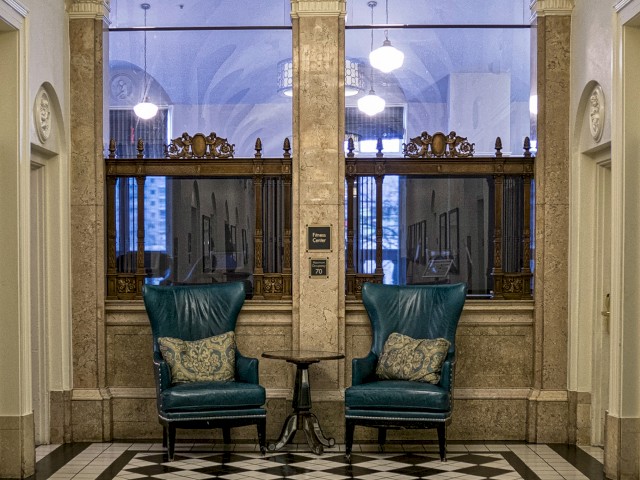 Elegantly decorated room with two green chairs, a small table, and checkerboard flooring, illuminated by a chandelier, and arched windows in the background.