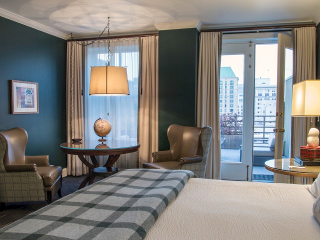 A cozy room with a bed, two armchairs, a round table, a globe, lamps, and large windows with city views.