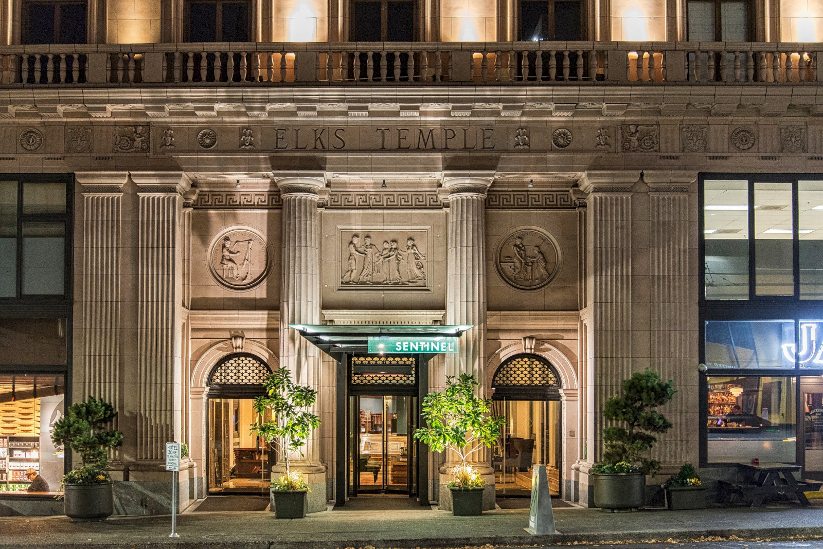The image shows the entrance to a grand, ornately decorated building at night. The sign above the entrance reads 