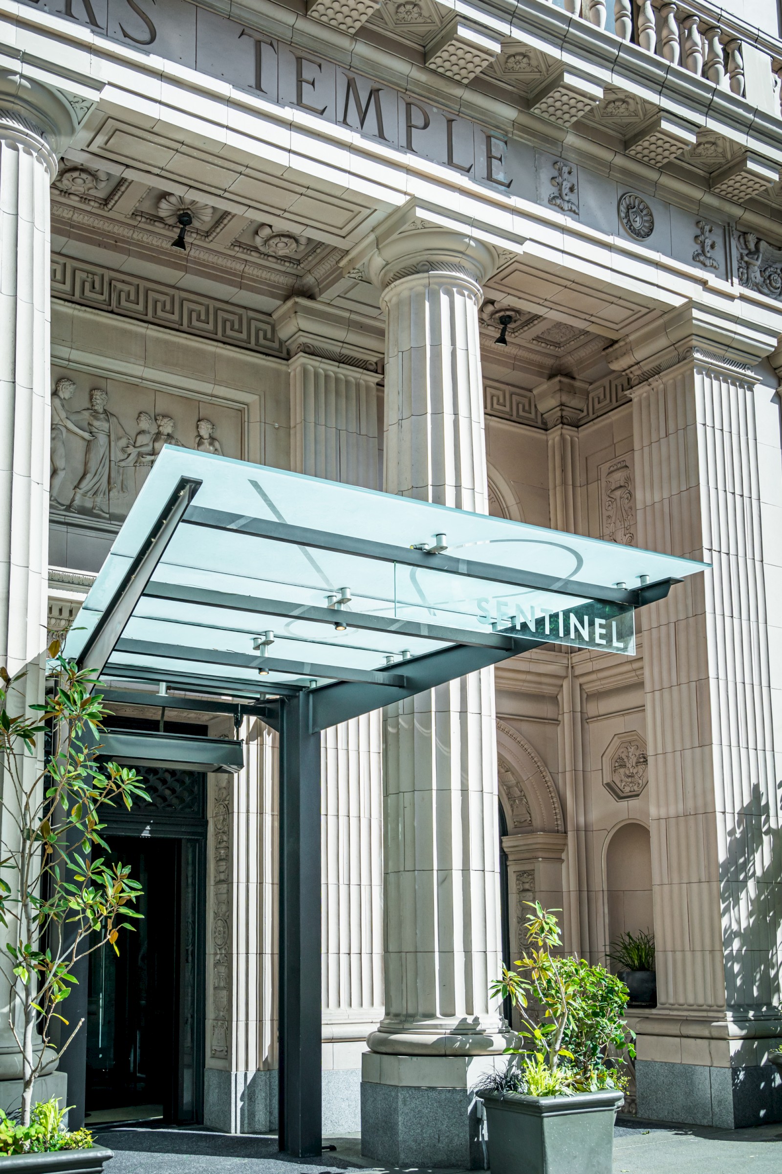 The image depicts an entrance to a building with classic architectural columns and a glass canopy, with plants in pots.