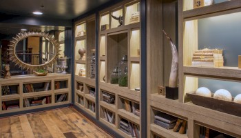 The image shows a well-lit room with wooden shelves filled with books, decorative items, and framed artifacts. A sunburst mirror is on one wall.