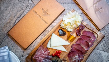 A charcuterie board with assorted meats, cheeses, and garnishes, accompanied by two folded menus on a wooden table.