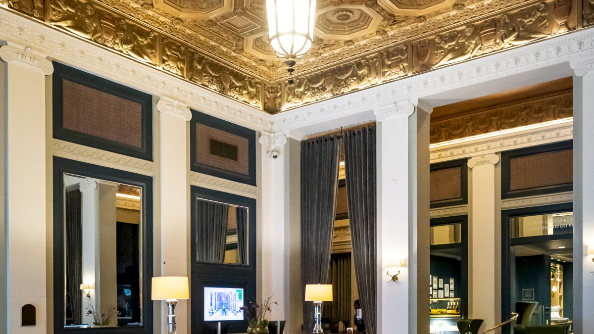 An elegant room with a high, ornate ceiling, modern furnishings, and large windows. The space features a bookshelf, couches, and a central chandelier.