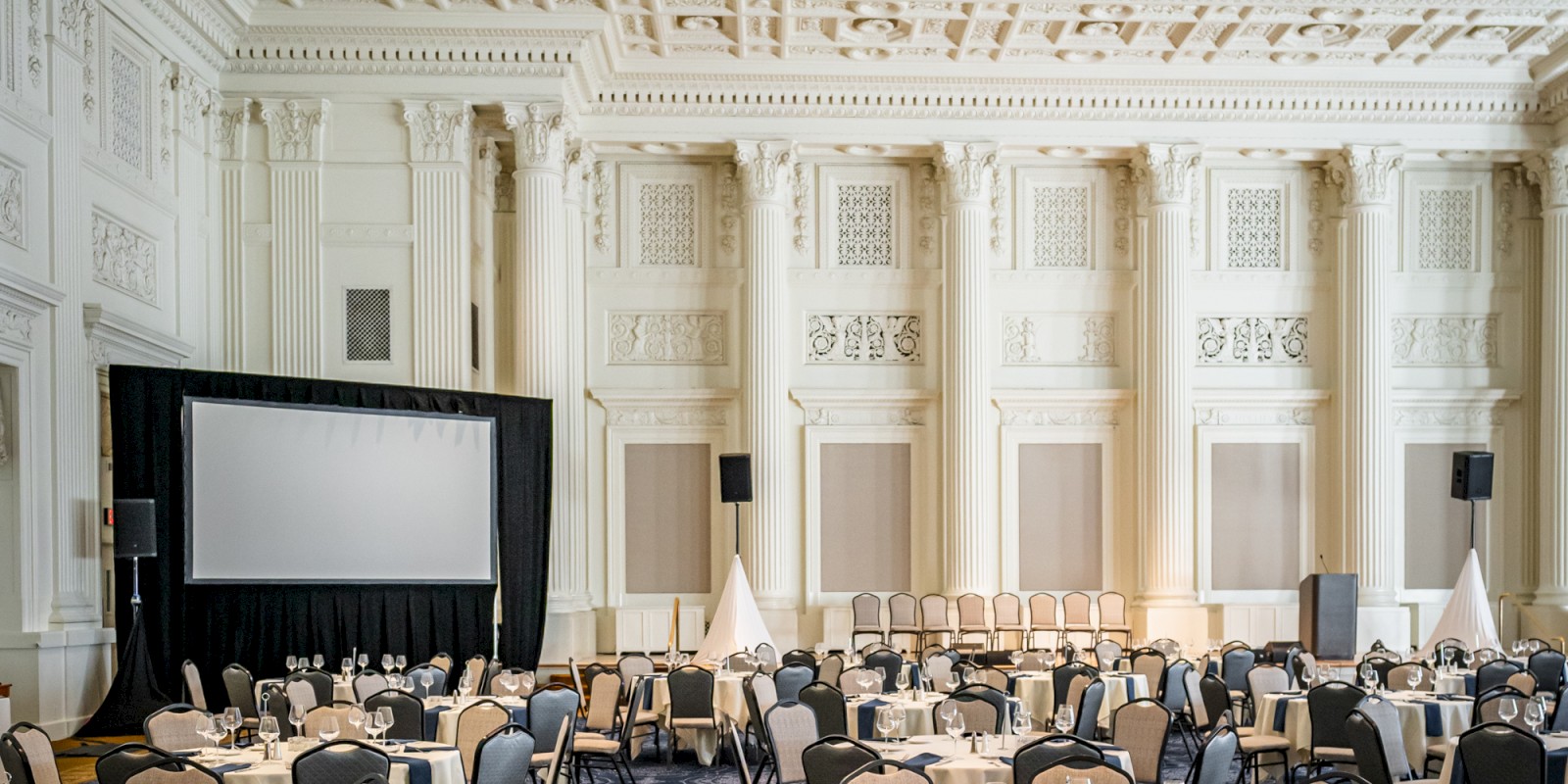 An elegant banquet hall with round tables and chairs set up, featuring ornate architectural details, a patterned blue carpet, and a large projection screen.