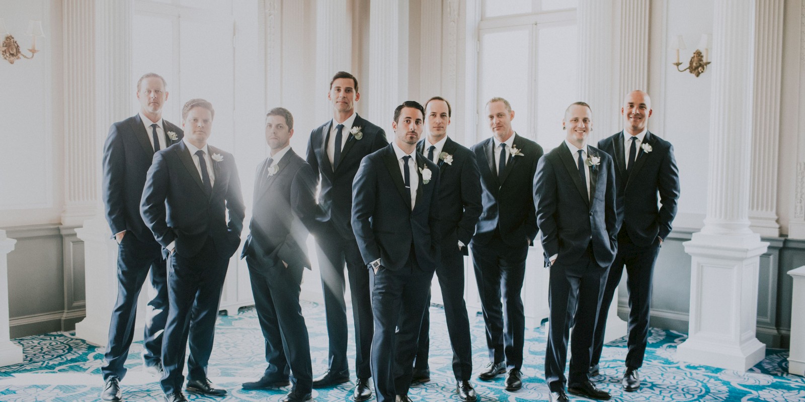 A group of nine men in suits are standing together in a formal setting with ornate decor and a blue-patterned floor.