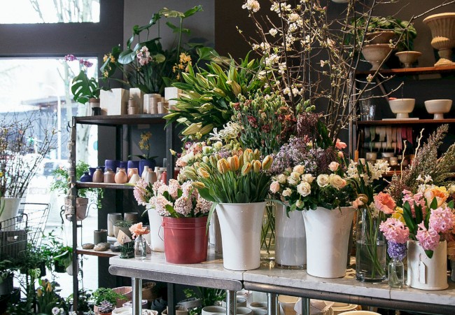 The image shows the interior of a flower shop, with an arrangement of various flowers in pots and vases on display tables.