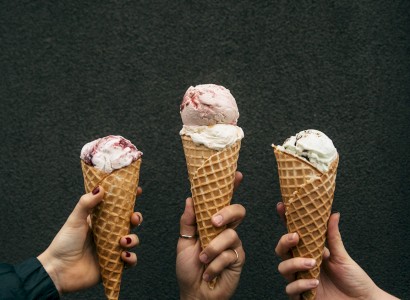 Three hands holding waffle cones filled with various flavors of ice cream against a dark background.
