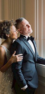 A couple, dressed elegantly, are standing near a window. The woman is in a white dress and the man in a black suit, both looking outside.