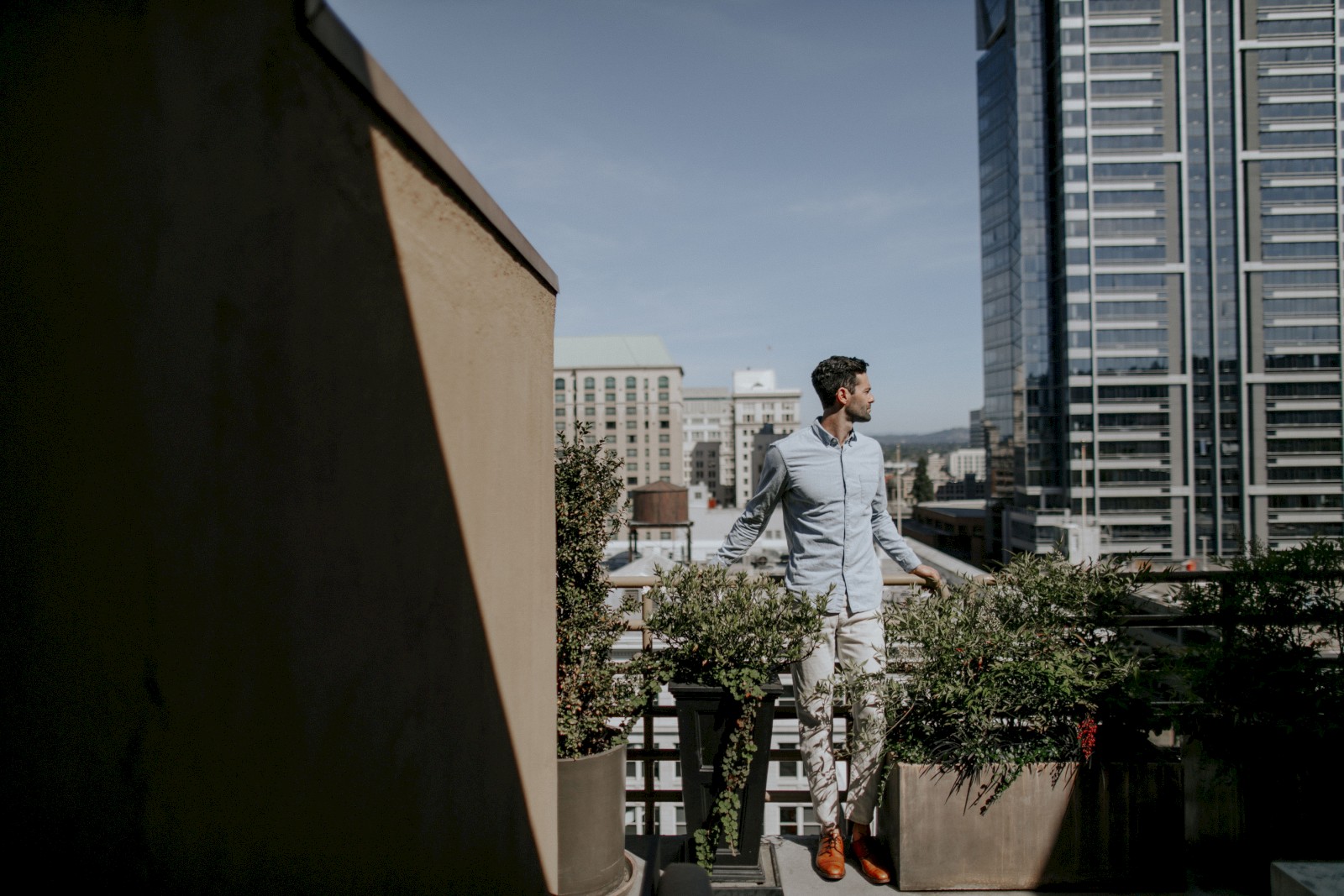 A man stands on a rooftop garden in a city, surrounded by potted plants and tall buildings, gazing off into the distance, possibly deep in thought.