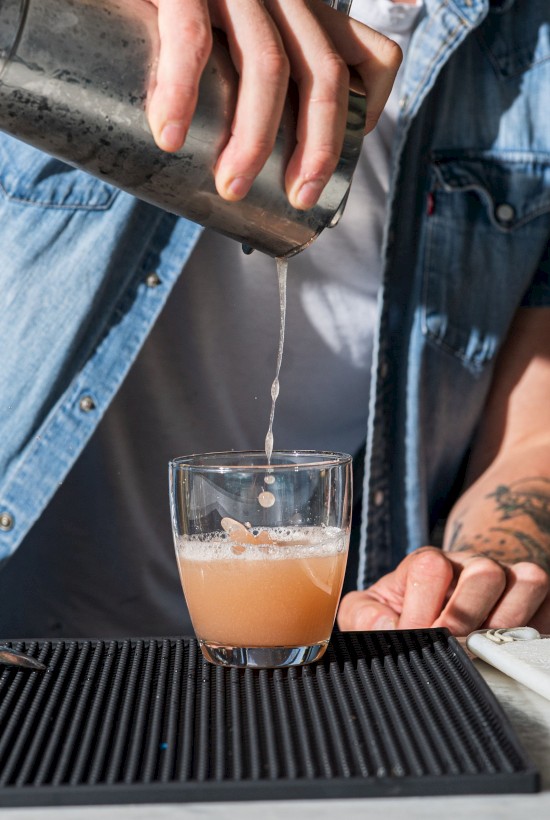 A person in a denim shirt is pouring a drink from a shaker into a glass placed on a bar mat.