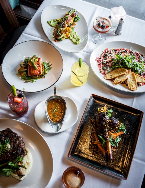 A table with various elegantly plated dishes, drinks, and condiments, featuring a mix of meat, pasta, salad, and vibrant cocktails.