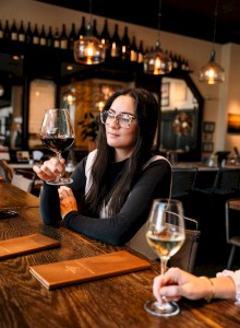 A woman is seated at a wooden table in a wine bar, holding a glass of red wine, surrounded by dim lighting and wine bottles.