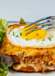 The image shows a person using a fork to cut into a croque madame with a fried egg, cheese, and ham, served with a side of leafy greens.