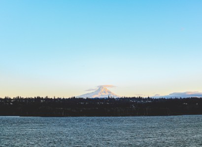 A distant snow-capped mountain under a blue sky, seen across a body of water with a treeline silhouette.