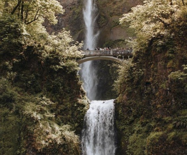 A two-tiered waterfall cascades down lush cliffs with a stone bridge spanning between the tiers, surrounded by greenery and trees.