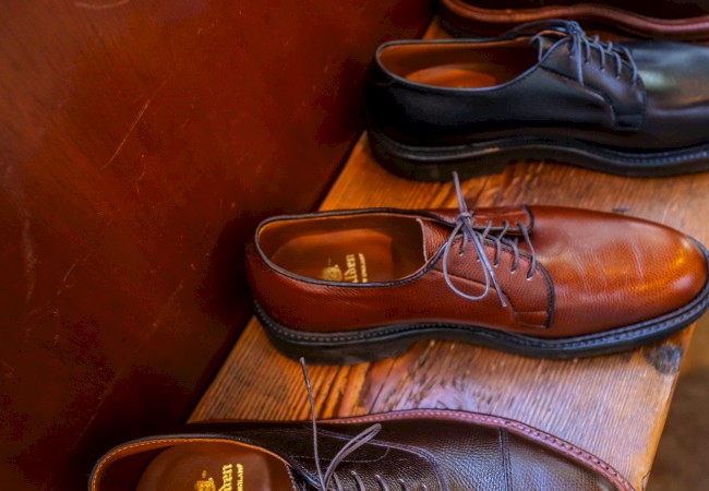 The image shows a row of leather dress shoes in various shades of brown and black, displayed on a wooden bench.