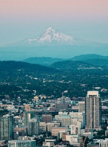 An urban skyline with various buildings in the foreground and a snow-capped mountain in the distant background under a gradient sky.