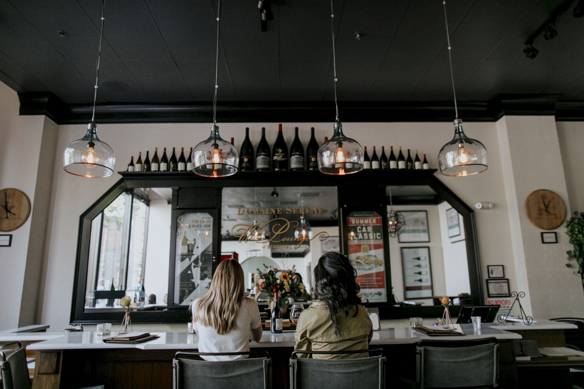 Two people are seated at a bar counter with hanging lights and decorative bottles. The atmosphere is cozy and modern, ending the sentence.