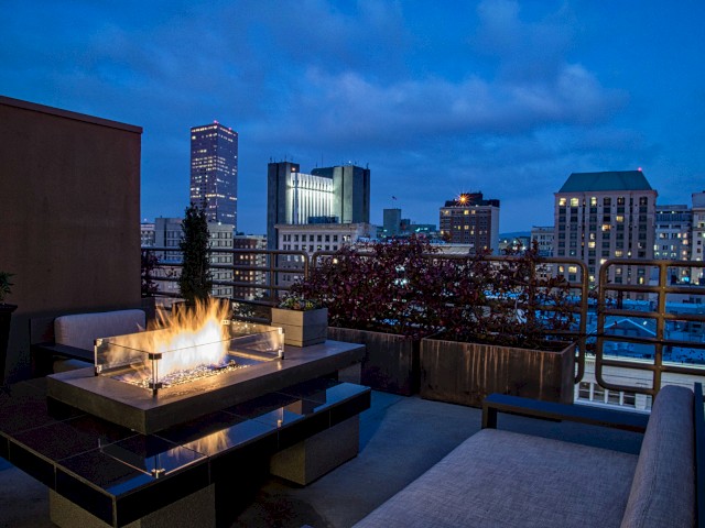 A rooftop patio with a fire pit, couches, and a view of a city skyline at dusk. The sky is a deep blue with scattered clouds.