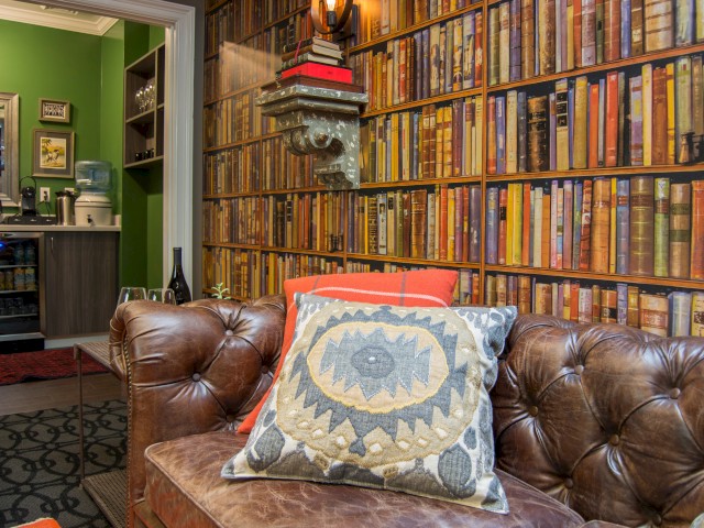 A cozy room with a leather sofa, decorative cushion, bookshelf wallpaper, and a view into a kitchenette with green walls in the background.
