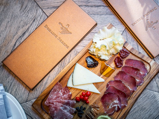 A charcuterie board with meats, cheeses, berries, and condiments sits next to two closed menus on a wooden table, with a folded napkin nearby.