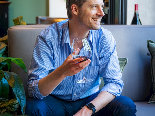 A man in a blue shirt is sitting on a sofa, holding a glass of wine and smiling, surrounded by plants and a table with a platter of food.