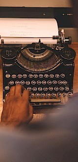 A person typing on a vintage typewriter with a sheet of paper inserted, viewed from over the shoulder on a wooden surface.