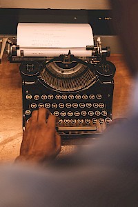 A person typing on a vintage typewriter with a sheet of paper inserted, viewed from over the shoulder on a wooden surface.