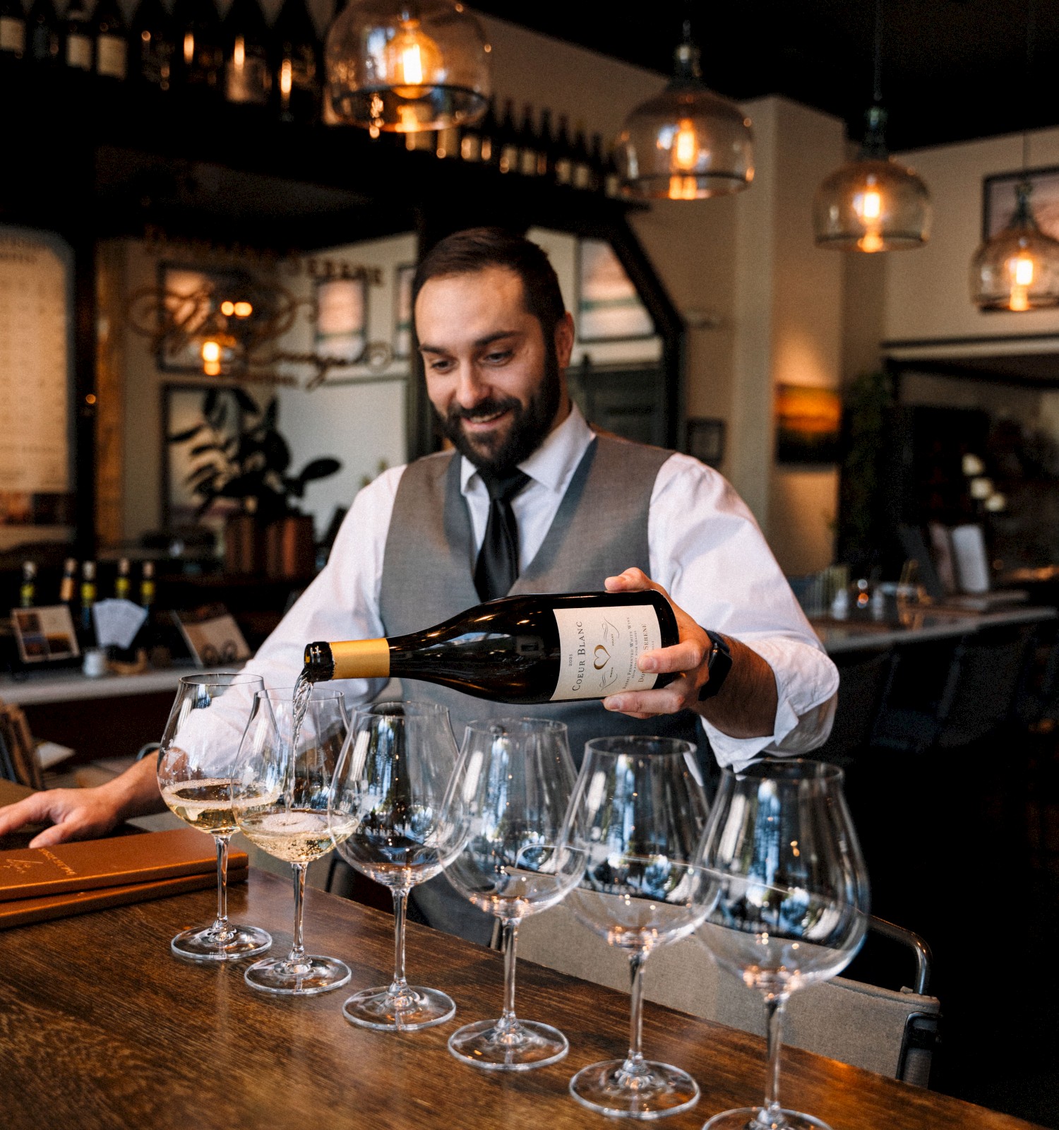 A bartender in a vest pours wine into several glasses lined up on a wooden bar counter, with hanging lights above in a cozy setting.