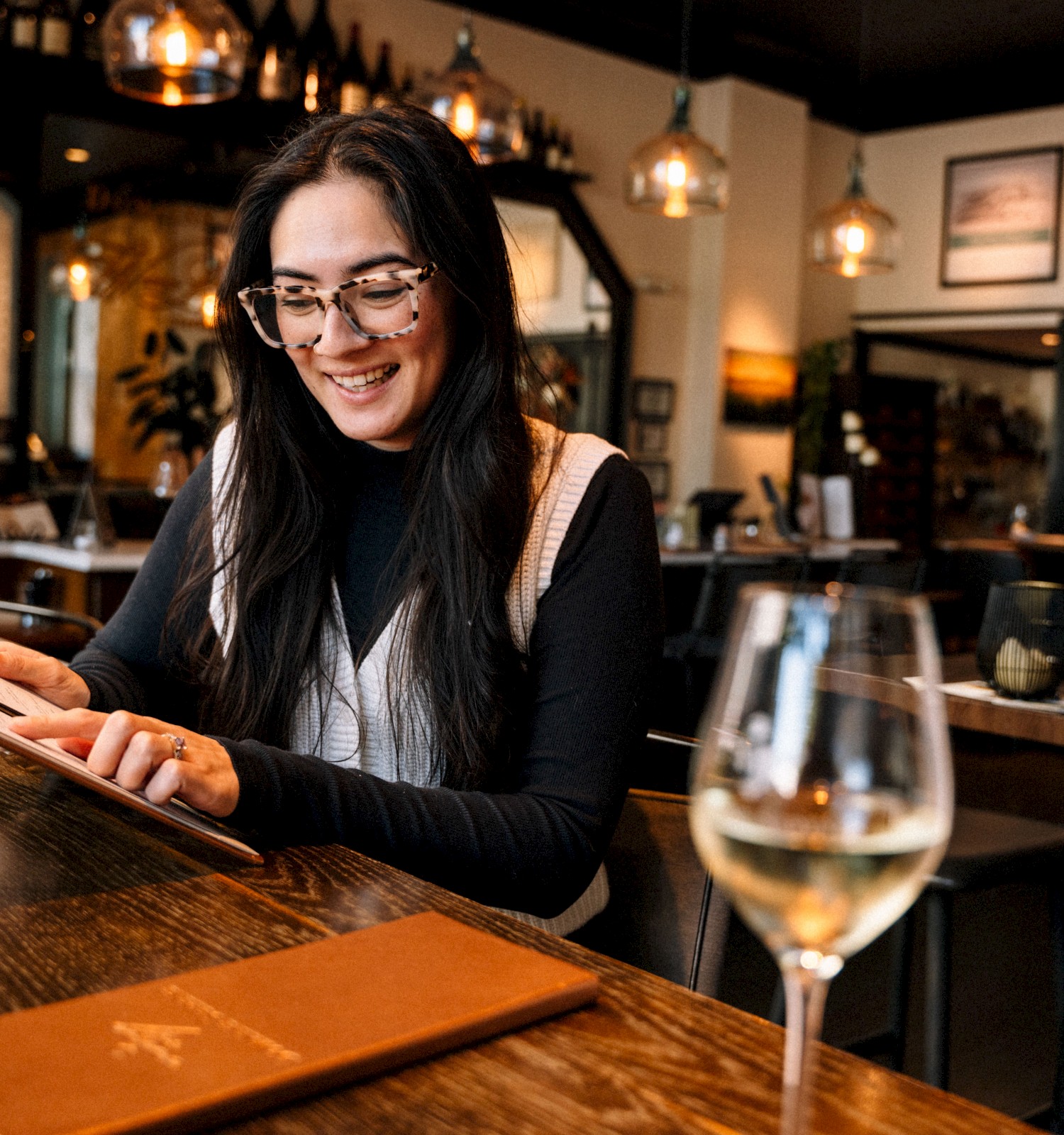 A person in a restaurant reads a menu, smiling. A glass of white wine is on the table. The setting is warm and inviting.