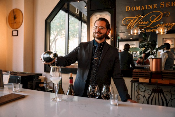 A person in a suit pours wine into a glass at a bar with bottles and glasses on the counter. The setting is a sophisticated wine lounge.