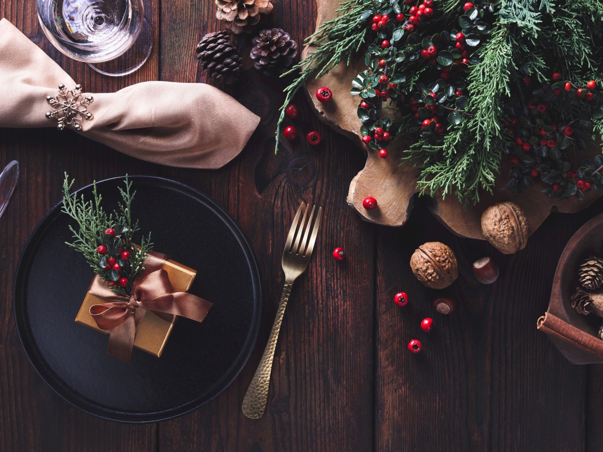 Festive table setting with a gift box and greenery on a plate, gold cutlery, nuts, and pine cones on a rustic wooden surface.