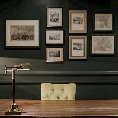 A vintage study room features a wooden desk, a brass desk lamp, a globe, and framed artwork on a dark wall.