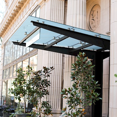 The image shows a building entrance with tall columns and a glass awning, flanked by potted plants along the sidewalk.