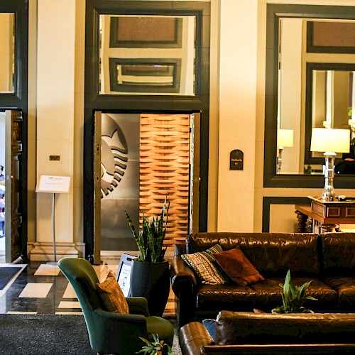 A cozy hotel lobby features leather sofas, mirrors, and decorative plants, with a person at the reception desk and an entrance door open.