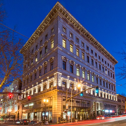 A large, illuminated building at night on a street corner, with light trails from traffic and trees nearby.