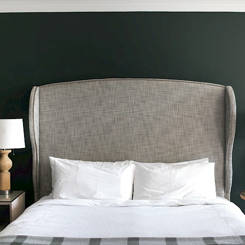 Deluxe King Room - A neatly made bed with a tall headboard, two pillows, and side tables with lamps, set against a dark wall.