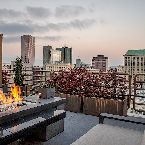 Terrace Studio Suite - A rooftop patio with a fire pit, seating, and city skyline views, surrounded by planters and decorative railings at sunset.