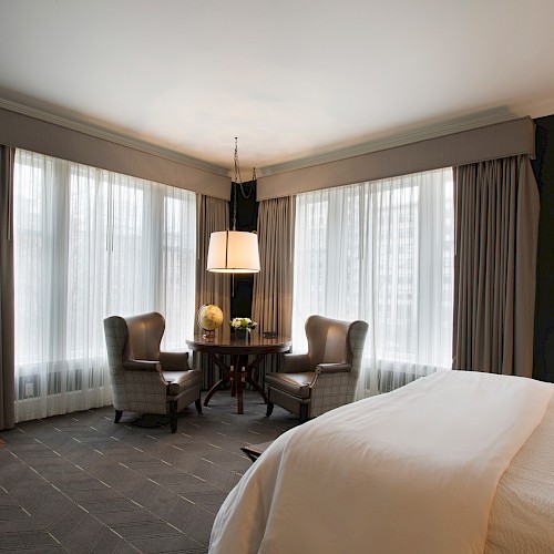 Premier Corner King Room - A cozy room with a large bed, two chairs by a window, a small table, and a fireplace, creating a warm and inviting atmosphere.