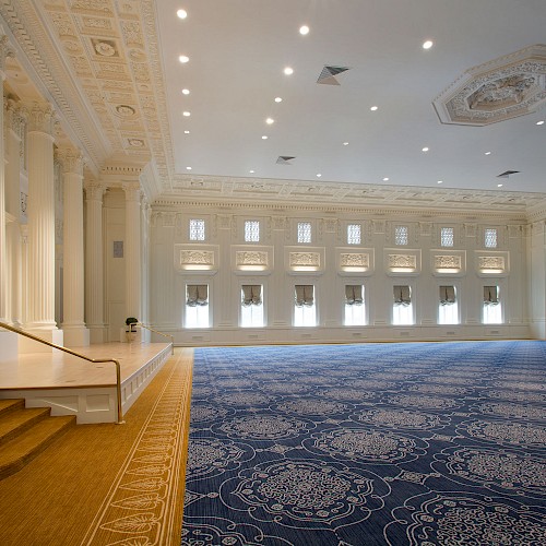 A spacious, elegant ballroom with ornate ceilings, columns, and a patterned carpet. Steps lead to a small stage with a potted plant.