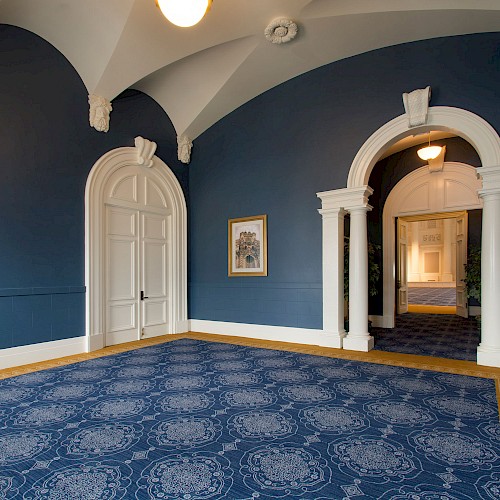 A room with blue walls, ornate arches, and a patterned carpet; a painting and a doorway to another area are visible.