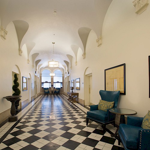 A long hallway with checkered floor, arched ceiling, blue chairs, framed pictures, and decorative plants at intervals.