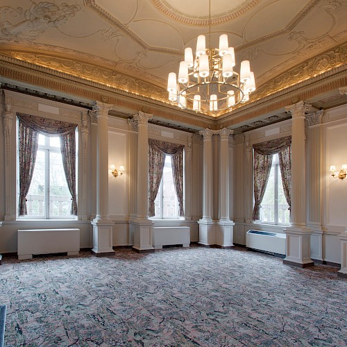 An elegant, ornate room with tall windows, columns, and a large chandelier, featuring floral curtains and decorative molding on the ceiling.