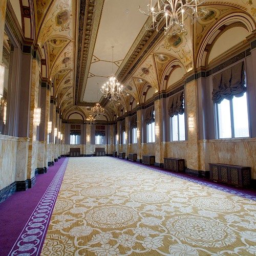 The image shows an elegant, spacious hallway with ornate chandeliers, arched ceilings, and lavish carpet, creating a grand, luxurious ambiance.